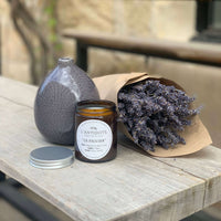 Duftkerze des französisschen Herstellers L'Antidote im Duft Le Figuier auf einem rustikalen Holztisch mit Lavendel. Die Kerze hat 150g und befindet sich in einem braunen Glas mit einem silbenen Schraubverschluss.