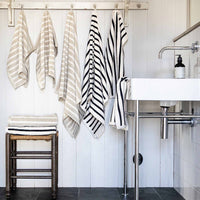 Badehandtuch 'Bonnie' von Tell Me More in Schwarz-Weiß gestreift, 70x140 cm, aus 100% Bio-Baumwolle – hängend drapiert in einem minimalistischen Badezimmer neben beigen Handtüchern und weißem Waschbecken
