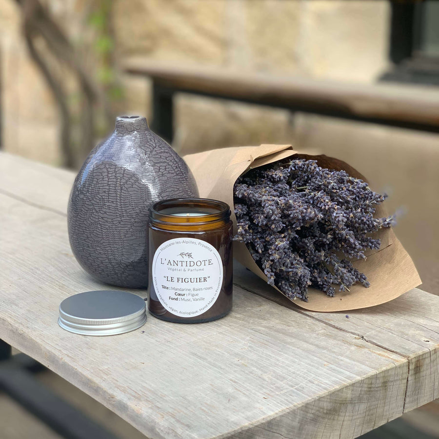 Duftkerze des französisschen Herstellers L'Antidote im Duft Le Figuier auf einem rustikalen Holztisch mit Lavendel. Die Kerze hat 150g und befindet sich in einem braunen Glas mit einem silbenen Schraubverschluss.