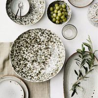 Stilvoll gedeckter Tisch mit handgefertigtem Keramikgeschirr in natürlichem Design – große Schale mit grün-weißer Sprenkelglasur, kleinere Schalen mit Oliven, Teller und Leinentuch, dazu Glas und Olivenzweig als Dekoration.