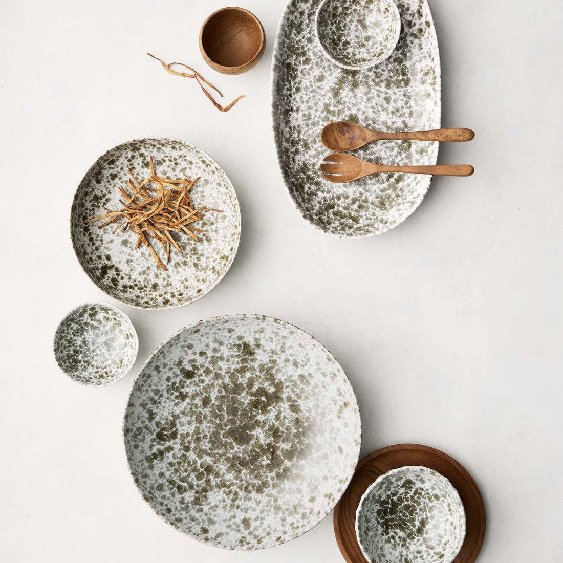 aServierschale ‘Rivoli’ von Tell Me More – Flatlay der Kollektion in verschiedenen Größen, weißes Porzellan mit olivgrünem Sprenkelmuster, Holzlöffel und Holzschälchen als Deko auf weißem Hintergrund