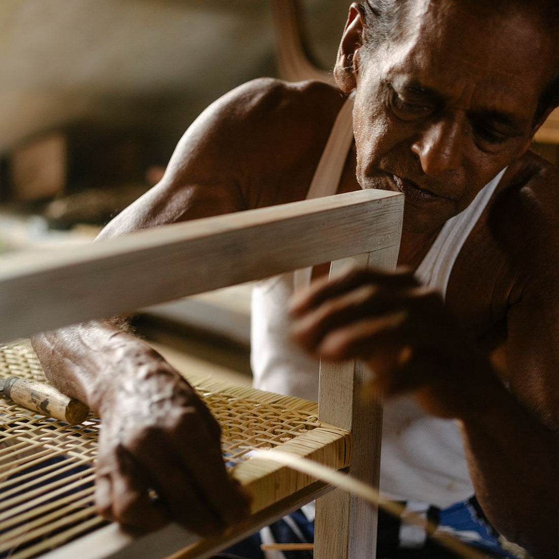 'Teppich 'Seegras Teppich mit Struktur' von Cane Cult aus Seegras, handgefertigt in traditioneller Handwerkskunst, Werkstattaufnahme eines Handwerkers bei der konzentrierten Flechtarbeit an einem Rattanstuhl'