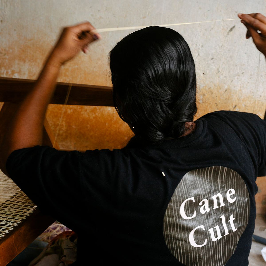 Leuchtenschirm 'Nymo' von Cane Cult aus geflochtenem Seegras in Naturfarben, boho-skandinavischer Stil, Werkstattaufnahme einer Handwerkerin bei der Flechtarbeit mit Cane Cult Logo auf dem T-Shirt