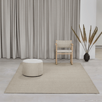 Teppich 'Escape Kelim' von Massimo aus Wolle in Hellbeige, natürlicher Stil, Lifestyle-Aufnahme in hellem Wohnzimmer mit Holzstuhl, rundem Pouf und Trockenblumen-Dekoration vor Leinenvorhängen, Nähte auf der Teppichoberfläche sichtbar
