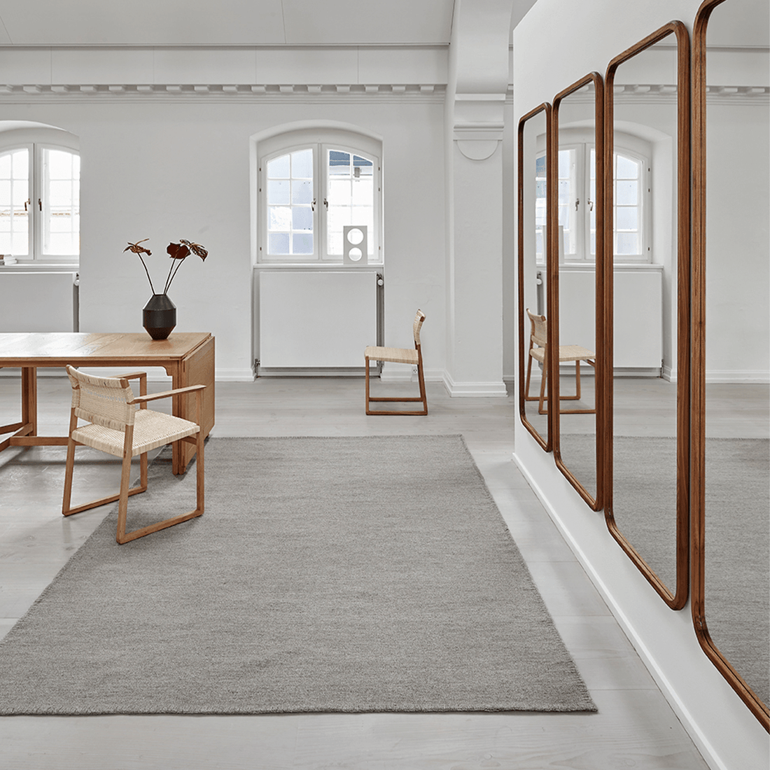 Teppich 'Escape Kelim' von Massimo aus Wolle in Steinfarben/Dunkelgrau, natürlicher Stil, Lifestyle-Aufnahme in hellem Loft-Wohnzimmer mit Holztisch, Rattanstühlen und großen Wandspiegeln, Nähte auf der Teppichoberfläche sichtbar