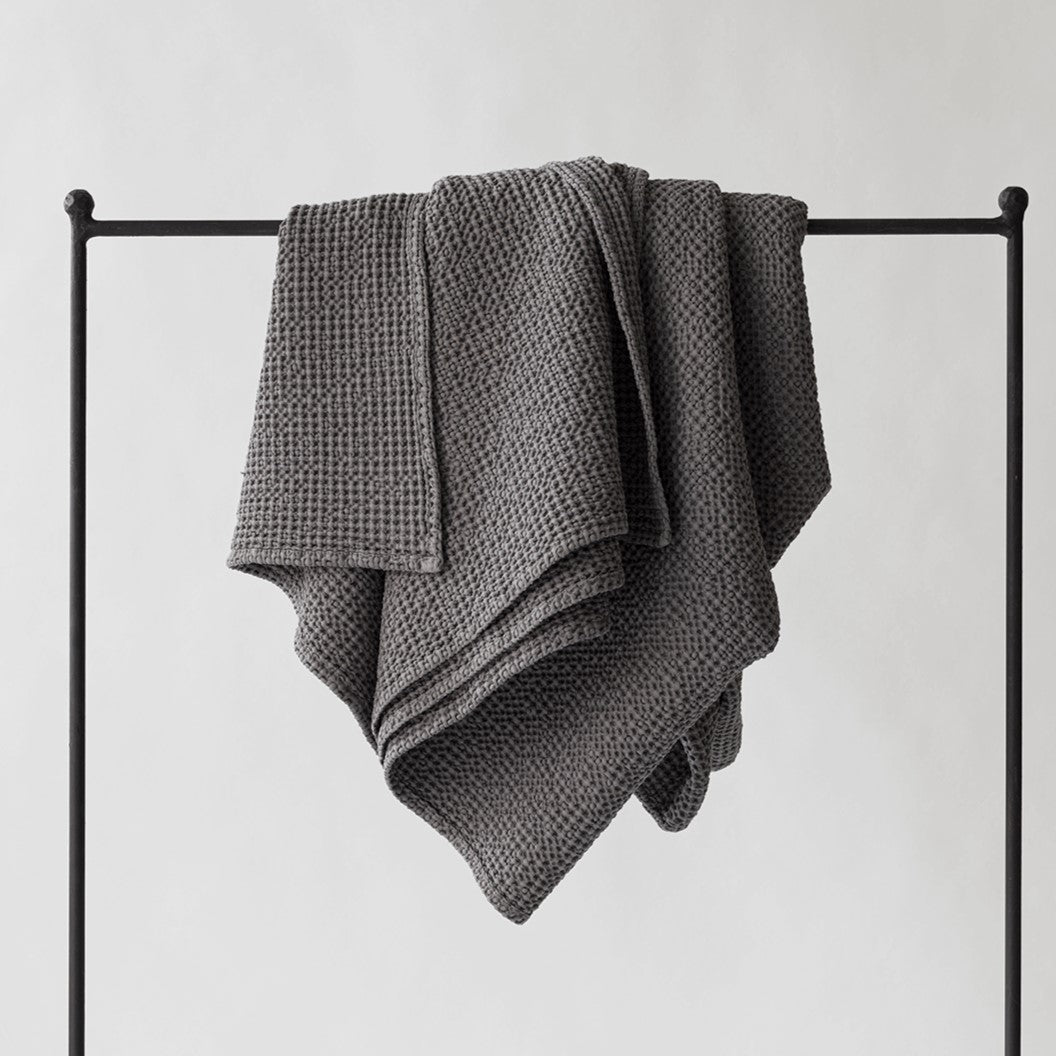 Decke 'Miro' dunkelgrau 140×200 cm von Tell Me More – Waffelpiqué-Decke über Kleiderständer drapiert, strukturierte Baumwolle, stimmungsvoller Schwarz-Weiß-Shot