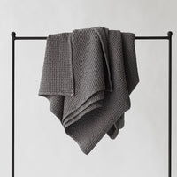 Decke 'Miro' dunkelgrau 140×200 cm von Tell Me More – Waffelpiqué-Decke über Kleiderständer drapiert, strukturierte Baumwolle, stimmungsvoller Schwarz-Weiß-Shot
